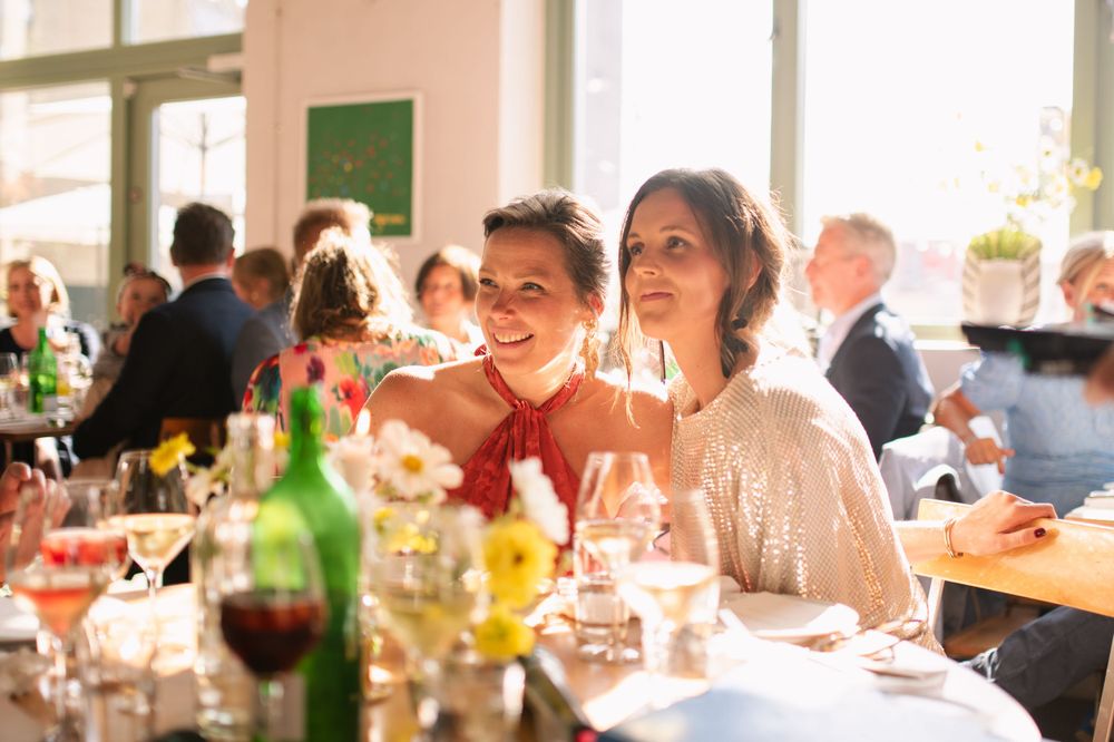 Wedding reception guests sharing drinks and laughter at a sunlit dining table.
