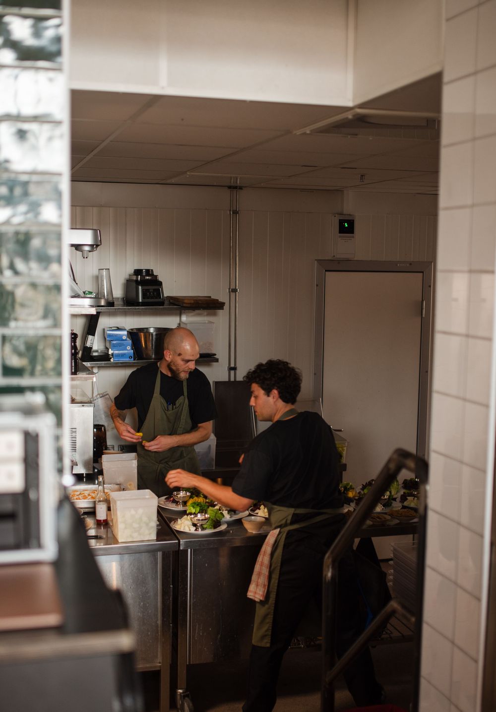 Two chefs preparing food in a restaurant kitchen with stainless steel prep tables and white tiled walls.
