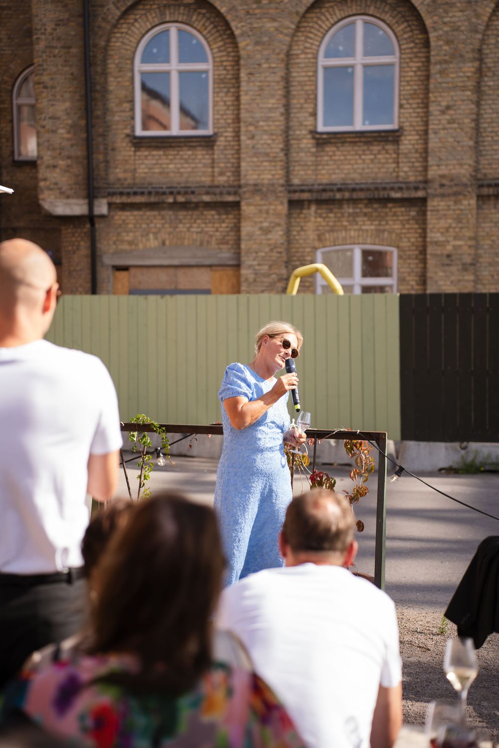 Someone in blue giving a speech at an outdoor event with brick building backdrop.