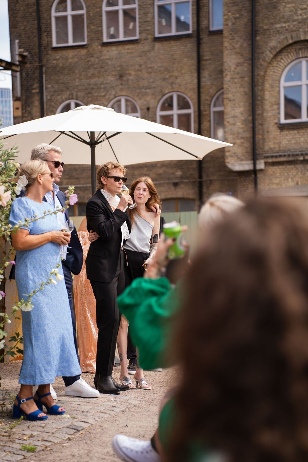 Wedding guests socializing under a white umbrella by a historic brick building.