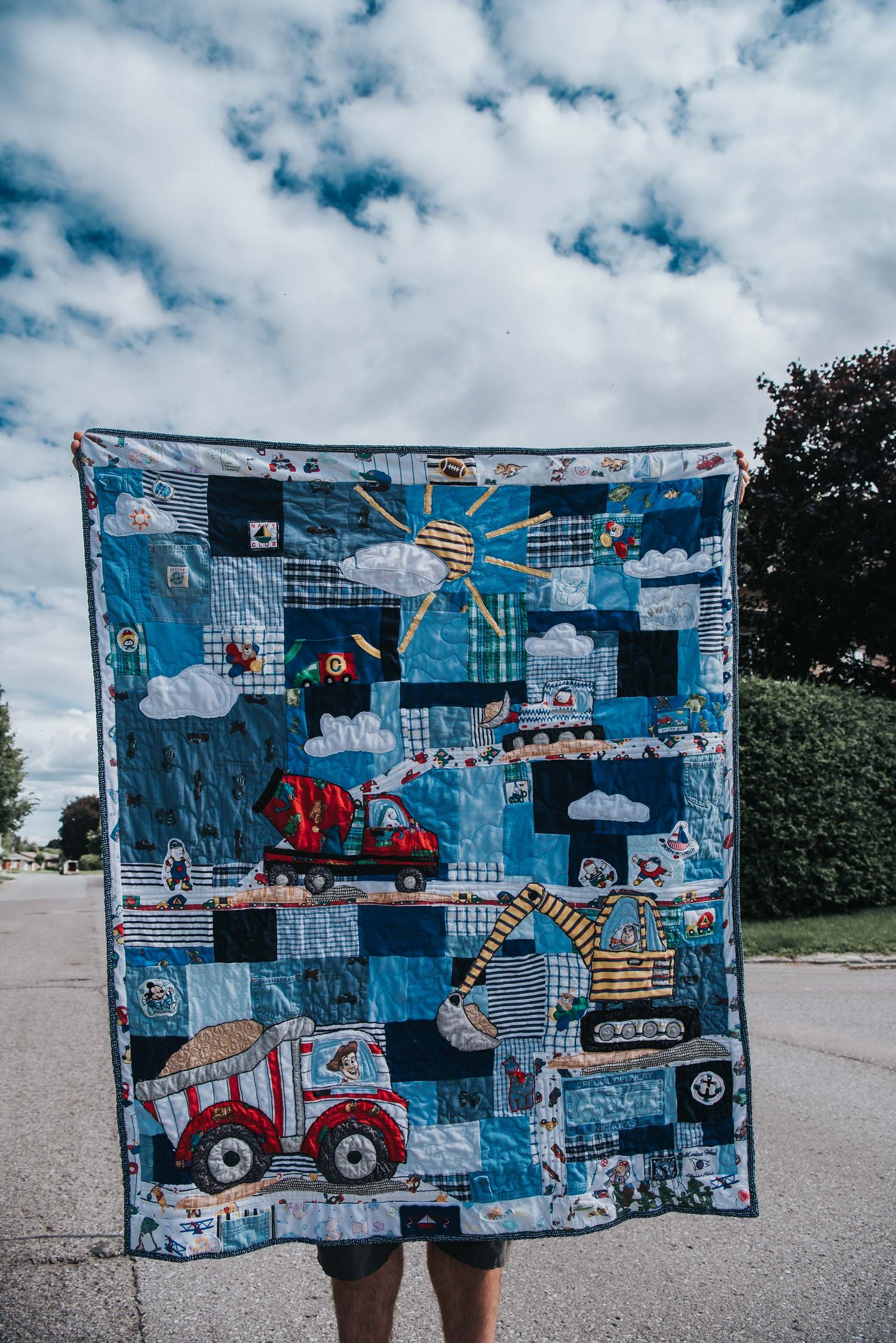 Construction themed memory quilt made with baby clothes and held up in front of a cloudy blue sky.