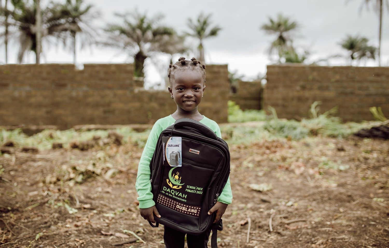 Young student wearing green shirt and backpack stands in rural setting with palm trees.