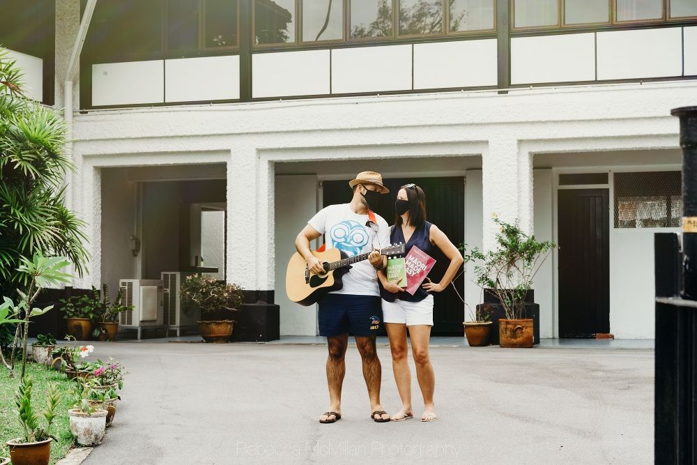 A couple outside their home in Singapore during lockdown. They are wearing masks, holding a guitar and language books.