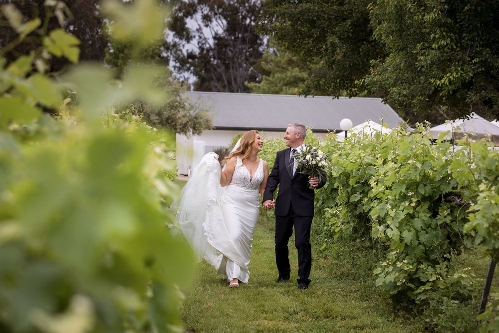 Bride and Groom walking holding hands and sharing a moment in the vines with the groom holding the wedding flowers