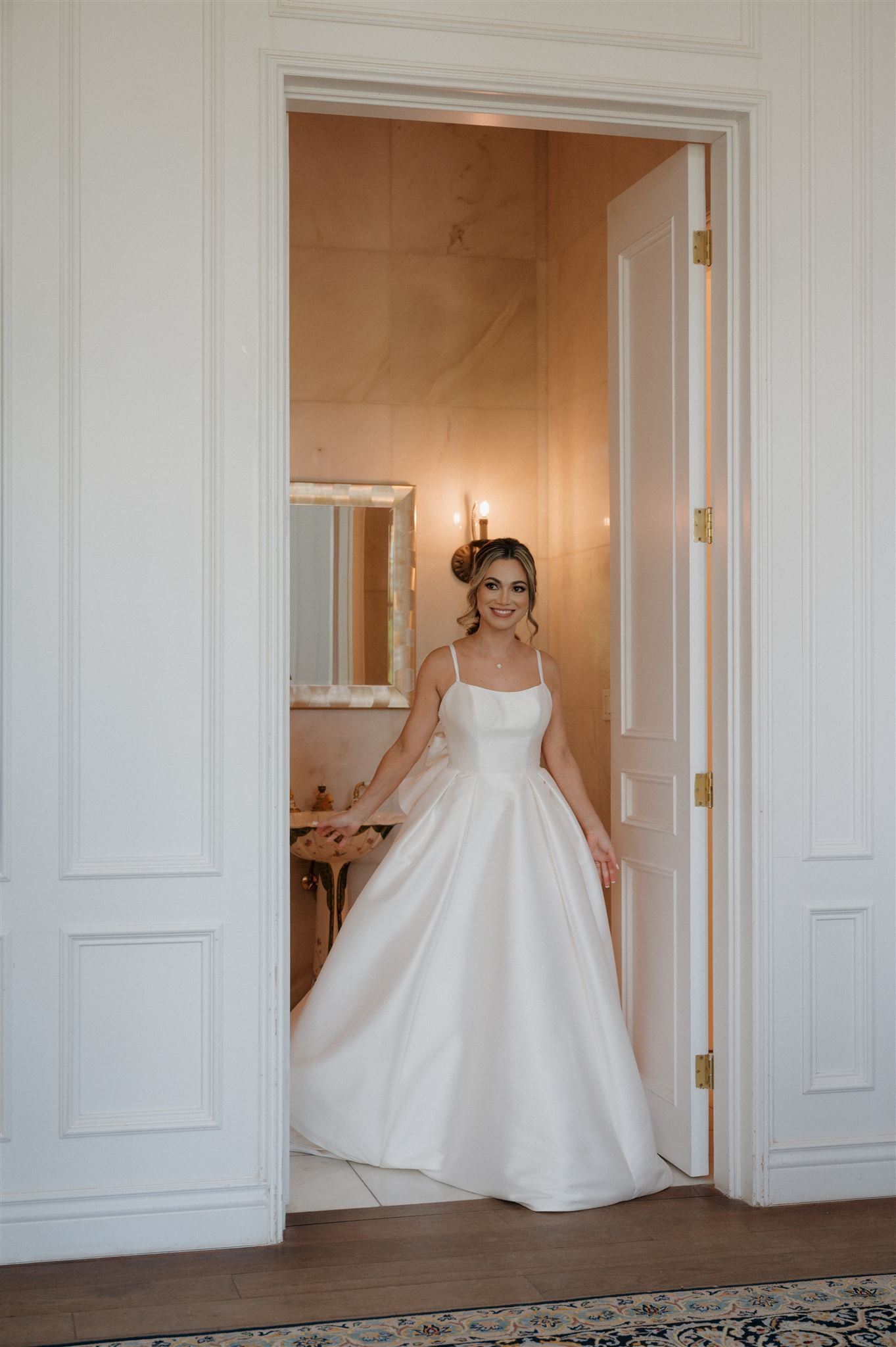 An elegant white wedding gown with a full satin skirt and spaghetti straps is shown in a doorway of a classic interior.