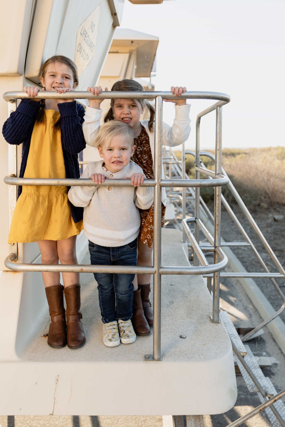 Three children smile while holding metal railing on an elevated outdoor platform near the coast.