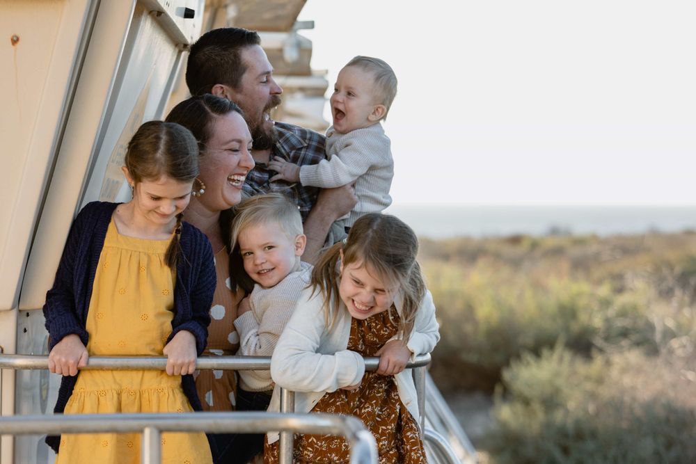 Laughing family with four children poses on a sunlit outdoor deck overlooking coastal landscape.