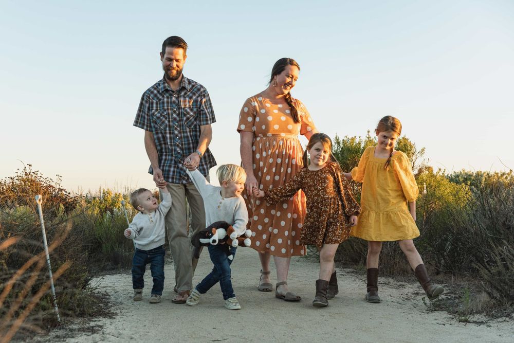 Pregnant mother and father walk with four young children on sandy coastal path at sunset.