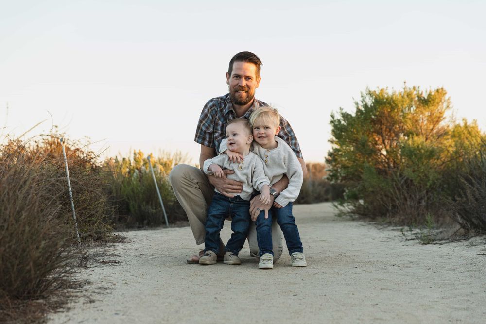 Father crouching on sandy path with two young children smiling outdoors at golden hour.