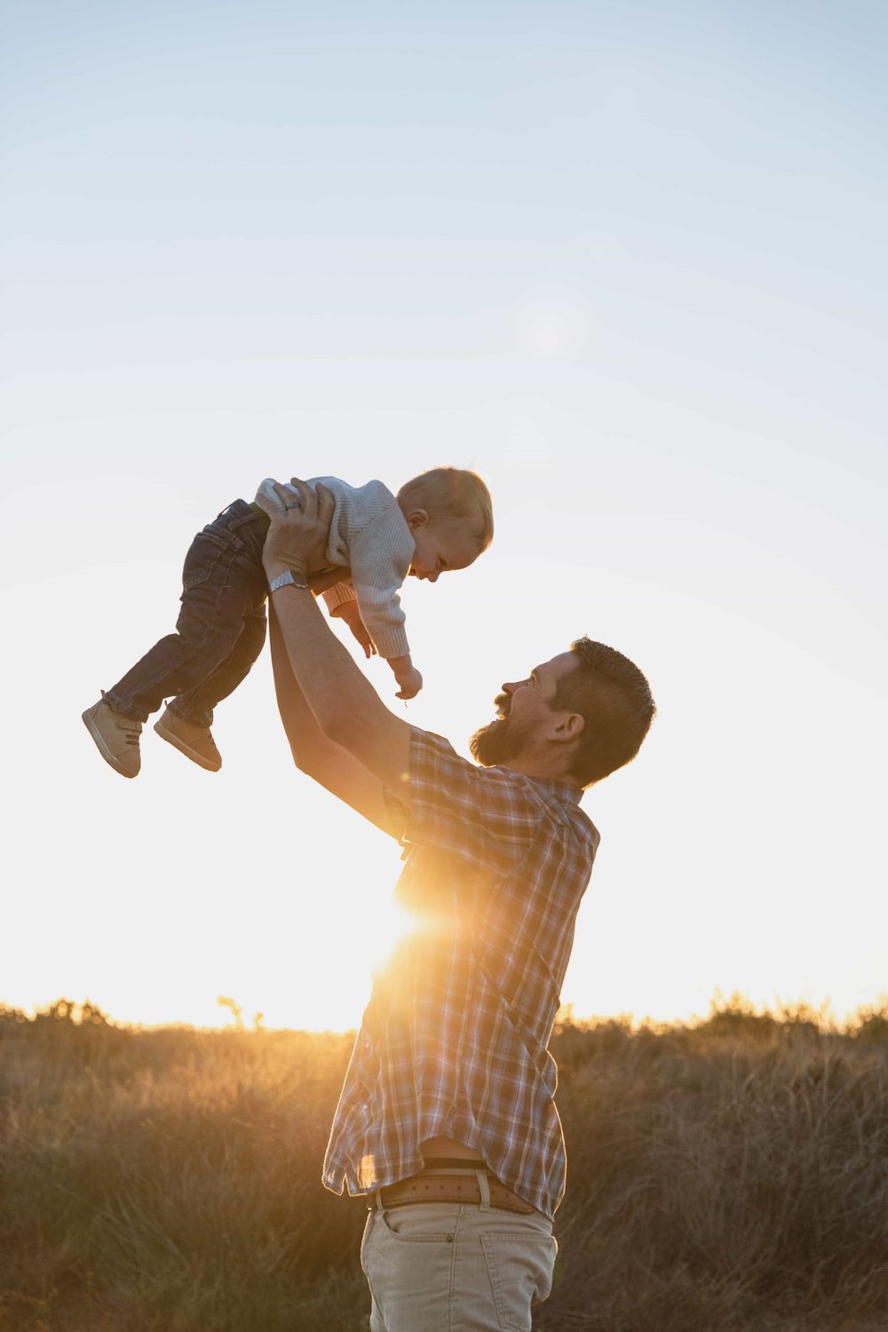 Father lifting baby joyfully into sunset sky in golden field at dusk.