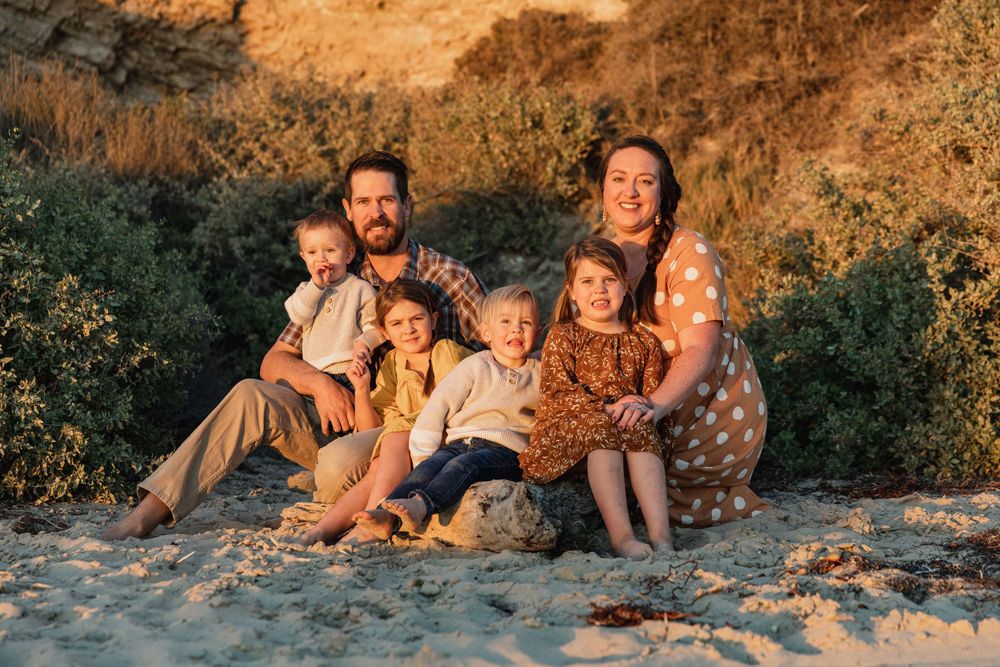 Family of six posing together on sandy beach surrounded by greenery at sunset.