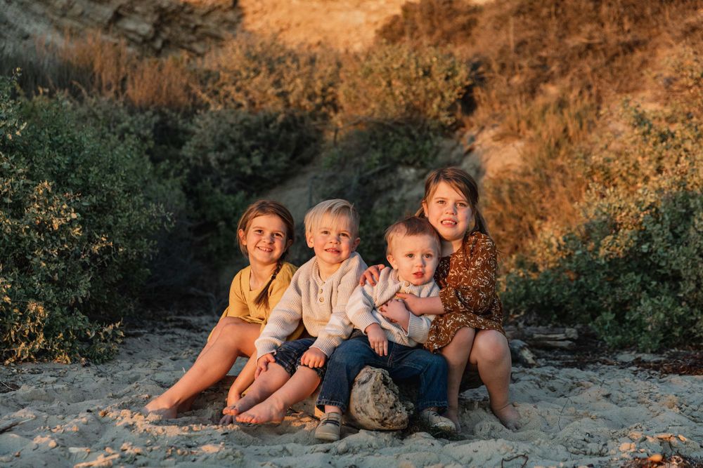 Four young siblings sitting together smiling on sandy beach at golden hour.