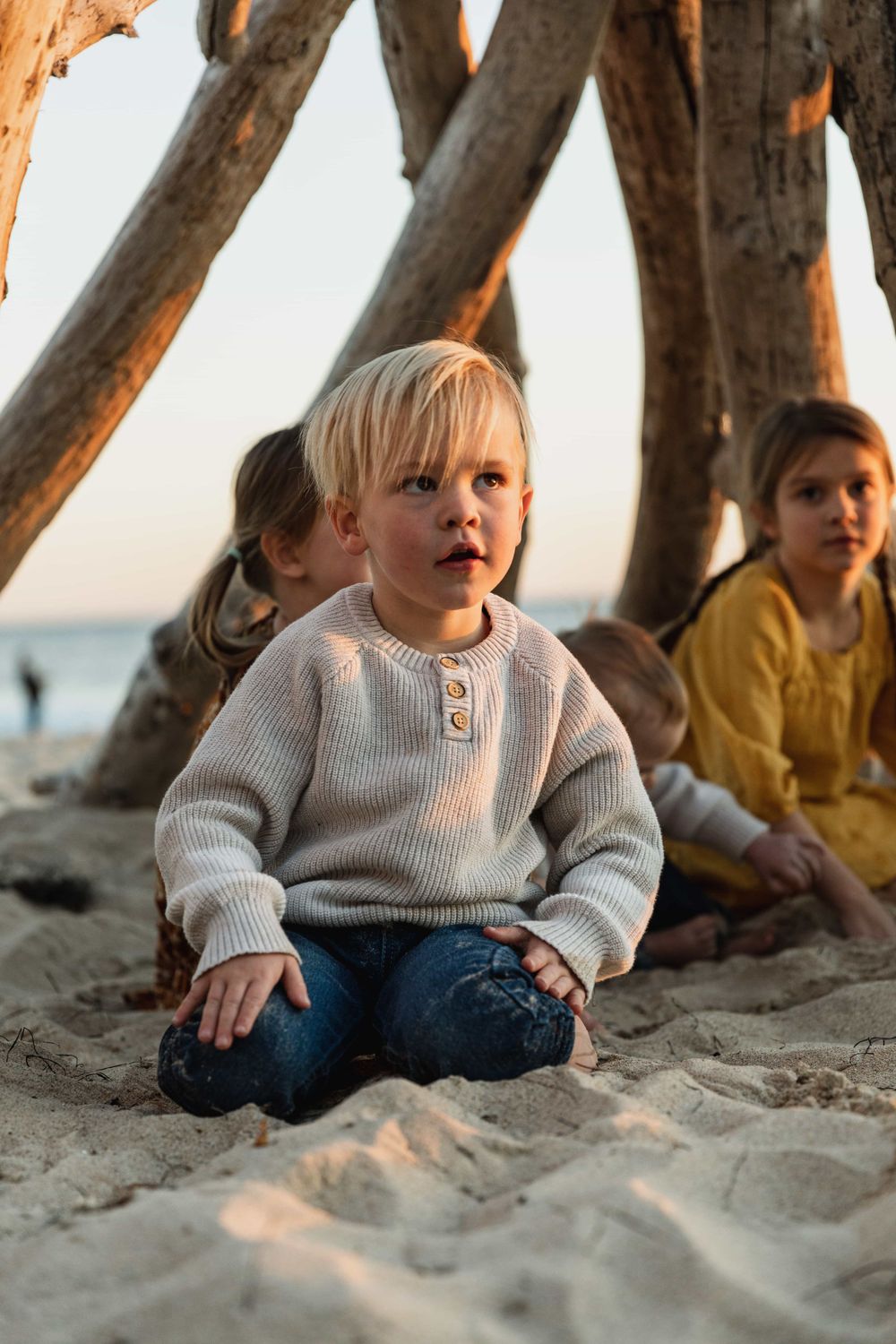 Blonde toddler boy sitting in sand under driftwood structure at beach sunset.