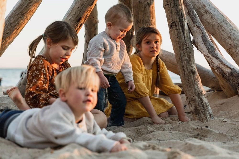 Four young children playing in sand under driftwood teepee at beach sunset.