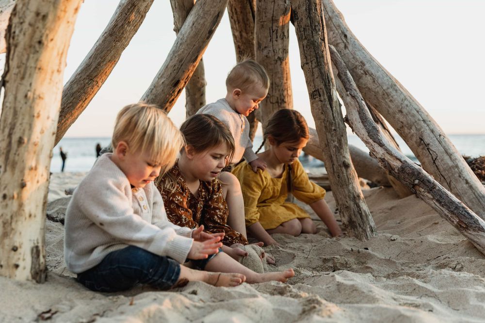 Four children playing together in sand under driftwood teepee at beach.