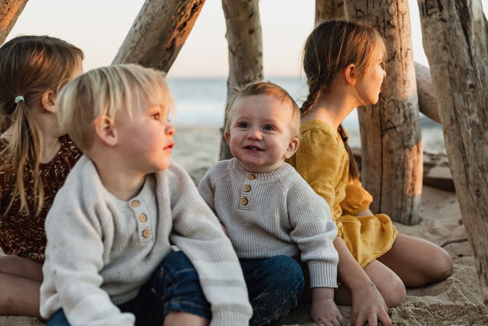 Three toddlers and a child sitting under driftwood structure smiling at beach.