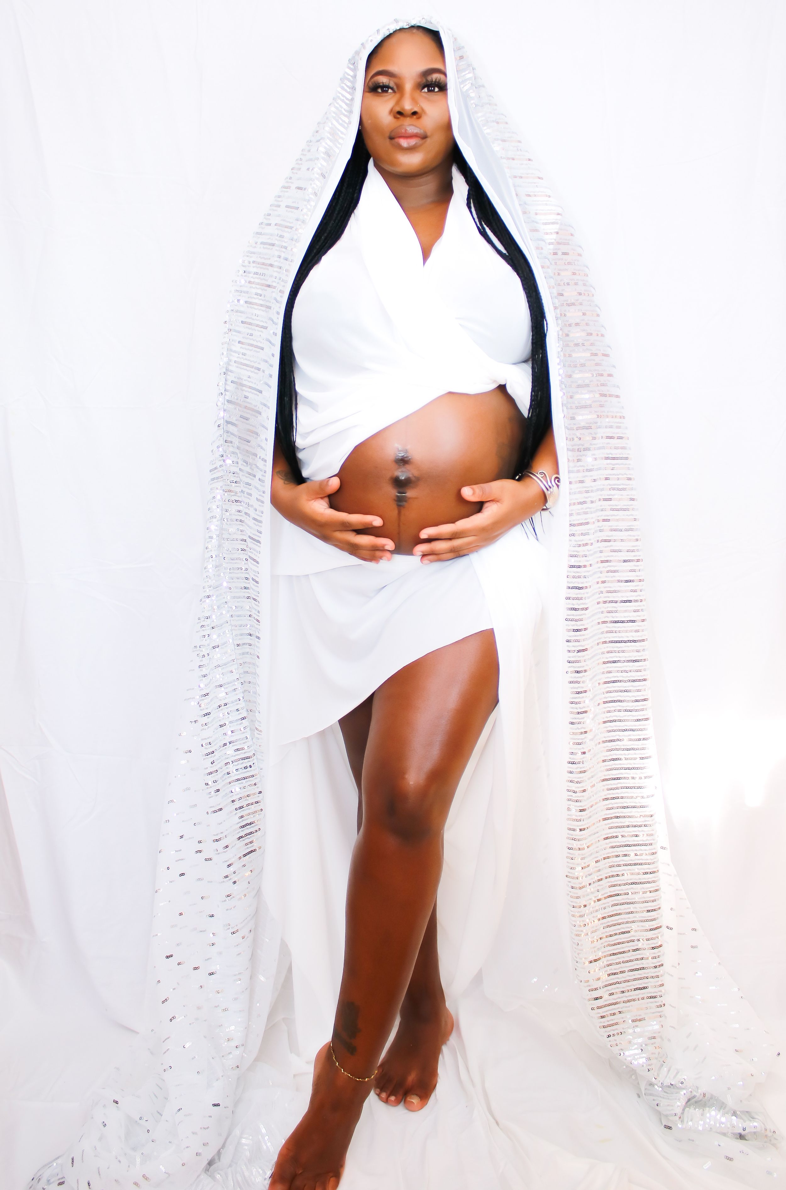 Elegant maternity photo with white wrap dress and flowing textured veil against light backdrop.
