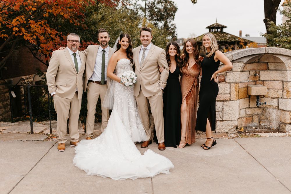 Wedding party of six poses outdoors in fall foliage, bride in lace gown with bouquet, groom in tan suit.