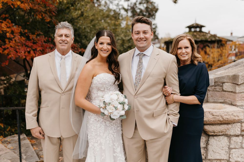 Bride and groom pose with parents outdoors, surrounded by vibrant autumn foliage at wedding venue.