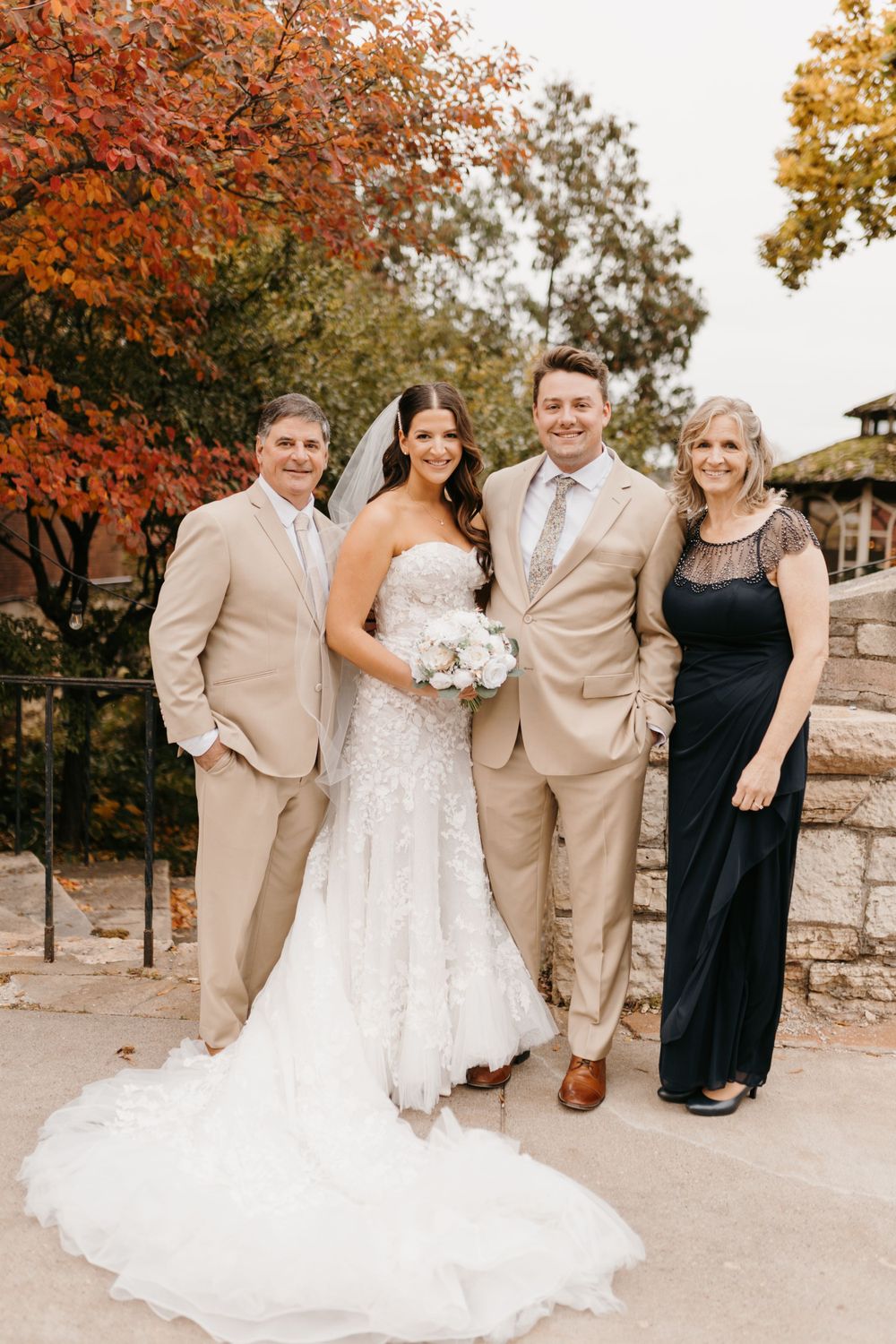 Bride in lace gown and groom in tan suit pose with parents before red autumn foliage.