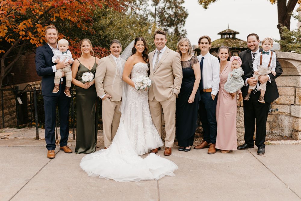 Large wedding party group photo outdoors with fall foliage, including children and extended family members.