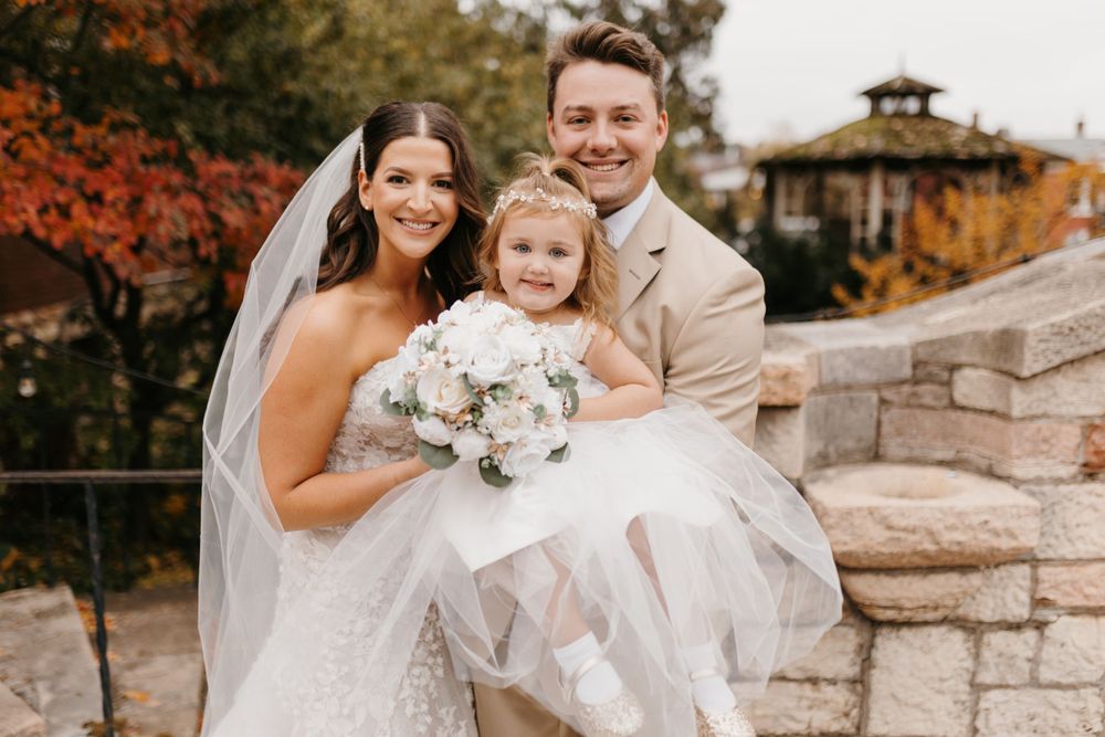 Bride and groom smile holding young flower girl in white dress outdoors at fall wedding.