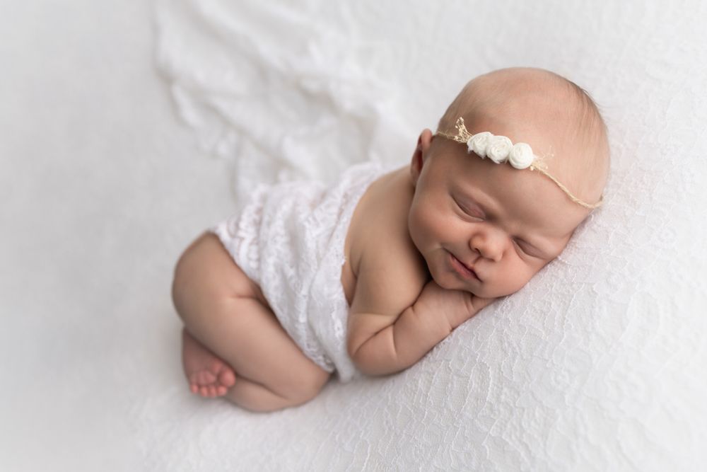 Newborn baby girl on white lace backdrop and white wrap around her chest smirking while sleeping.