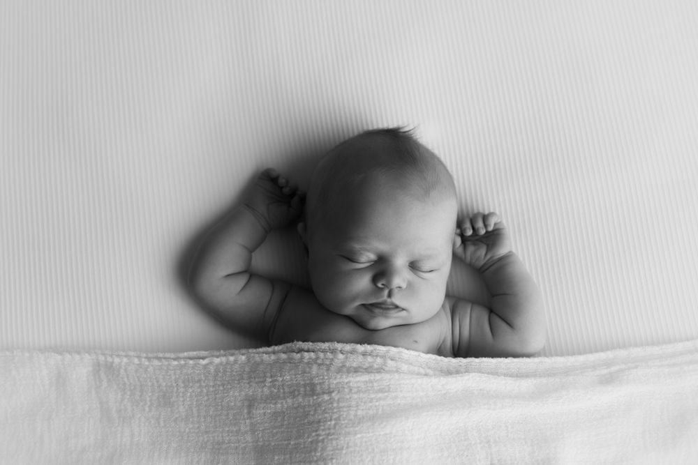 Black and white image of newborn baby girl laying on her back sleeping with her arms above her head.