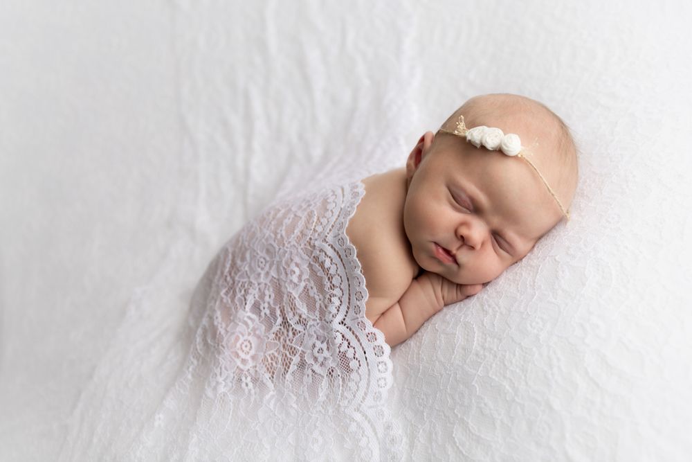 Newborn baby girl laying on white lace with white lace covering her back and white floral headband.
