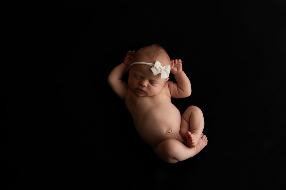 Newborn baby girl on black backdrop and white bow headband in a womb-like pose.