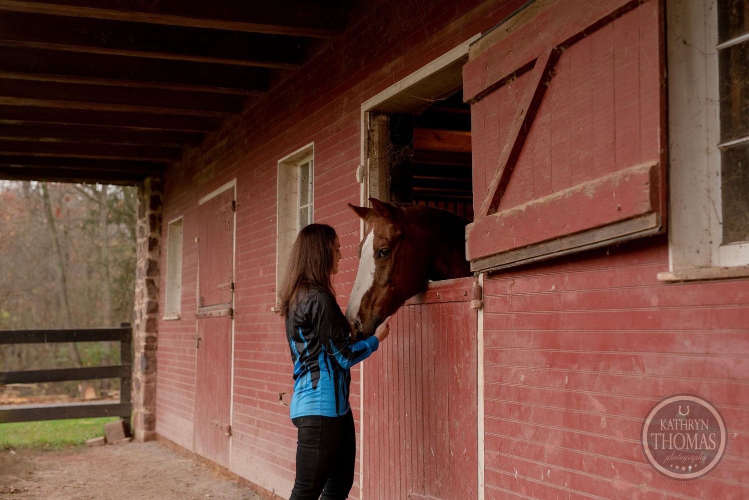 A horse peeks out from a rustic red barn stall while someone opens the door.