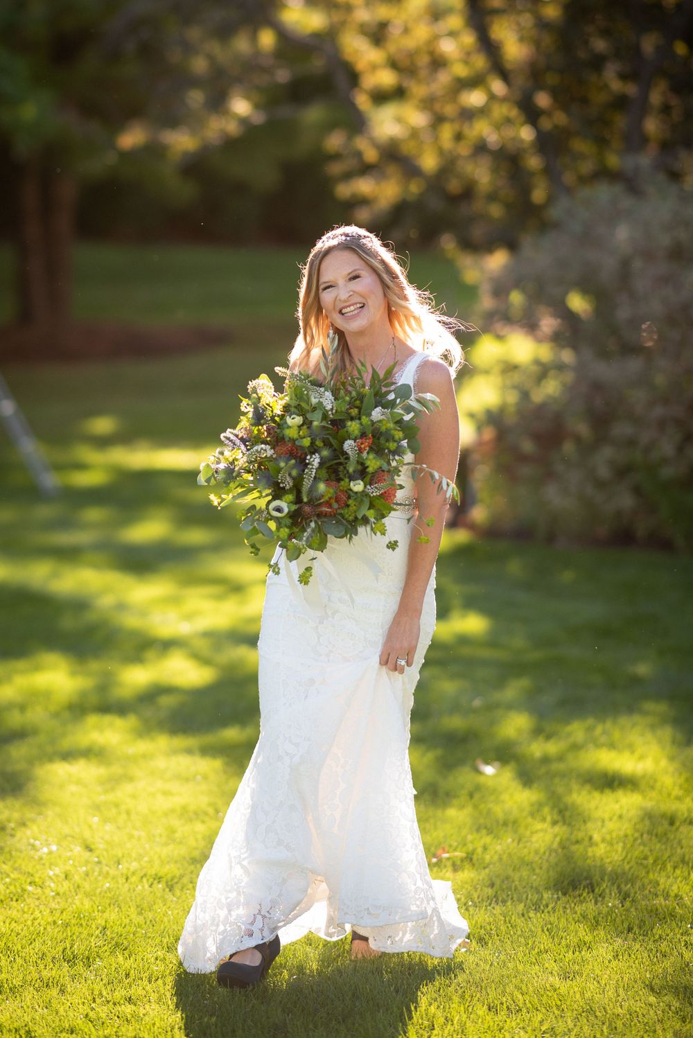 Bride in white lace dress picks flowers in a sunlit garden during golden hour.