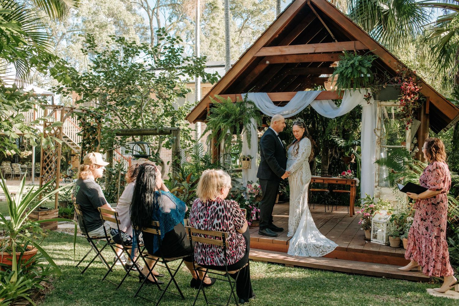 A wedding ceremony in a rustic garden gazebo with guests seated on chairs.