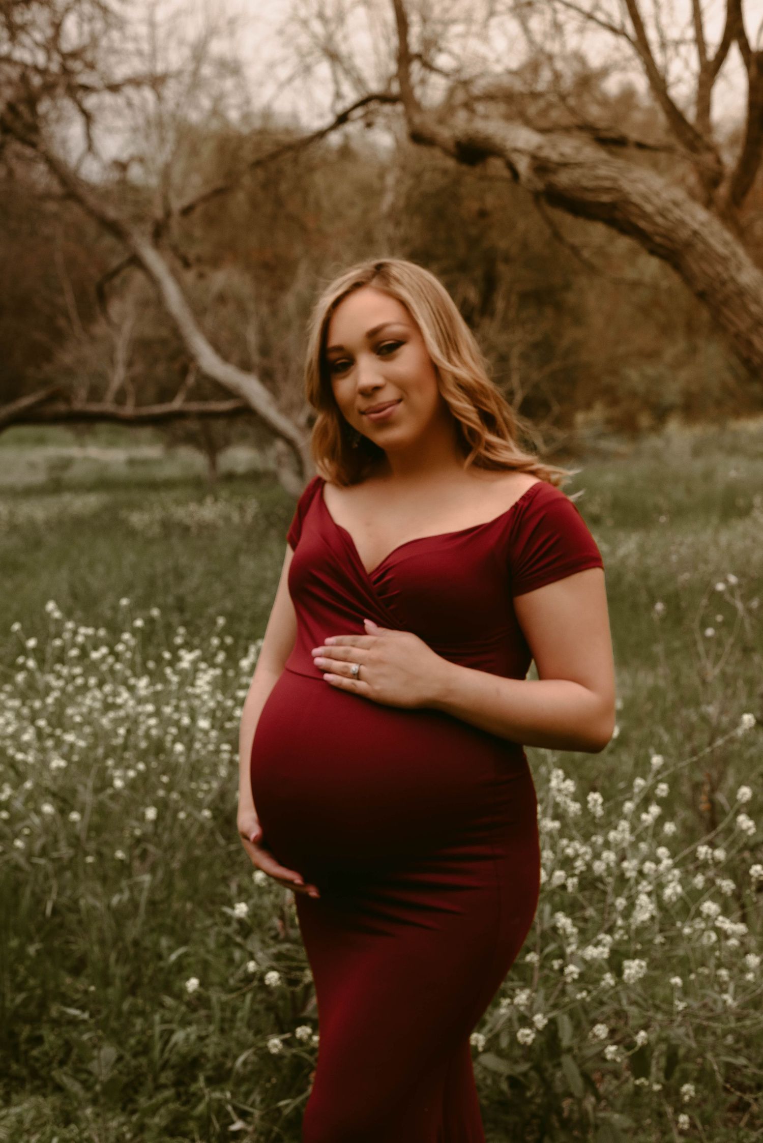 Pregnant woman in burgundy dress tenderly holds her belly in a natural outdoor setting.