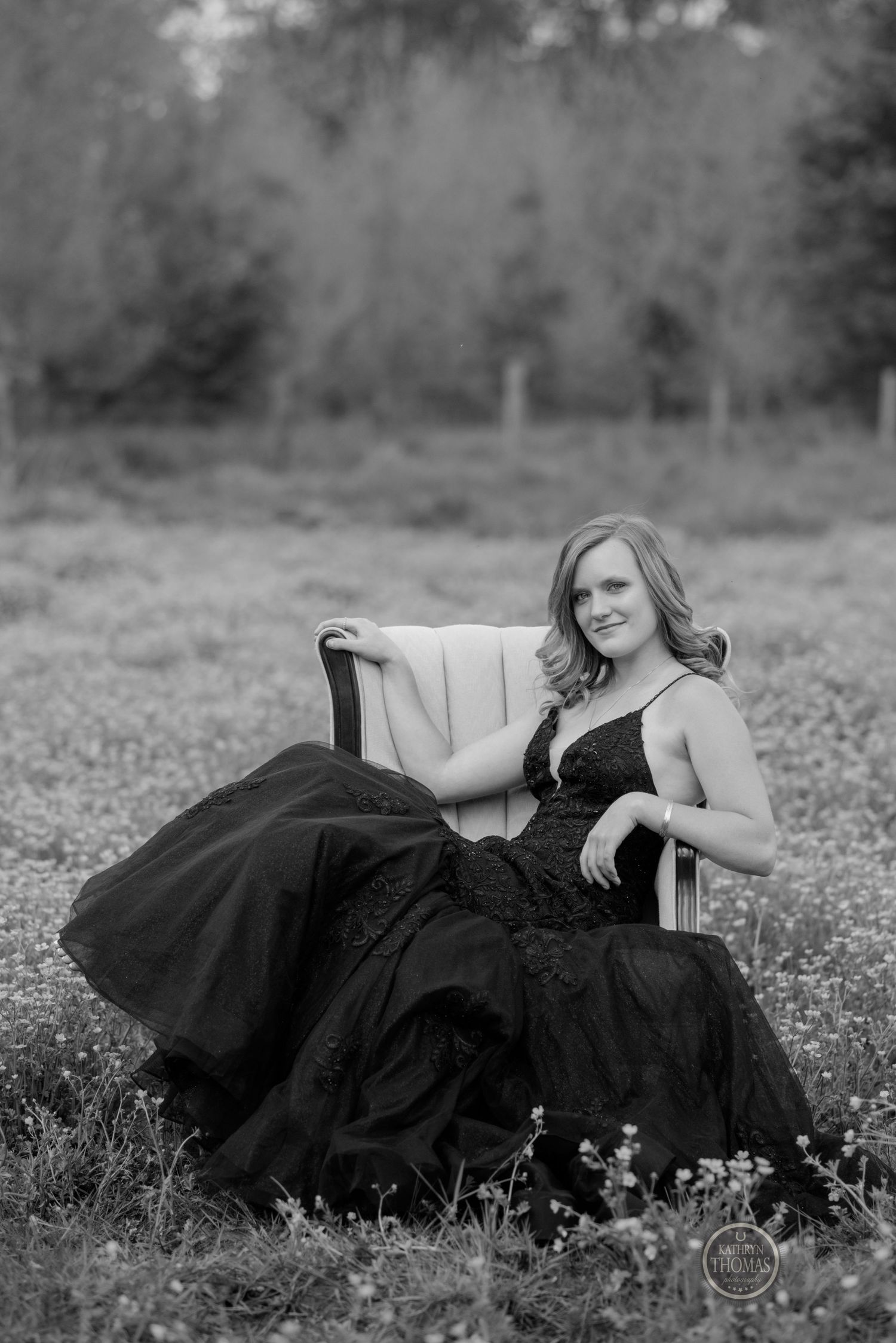 A black and white photo captures a figure in a formal gown seated in an armchair among wildflowers.
