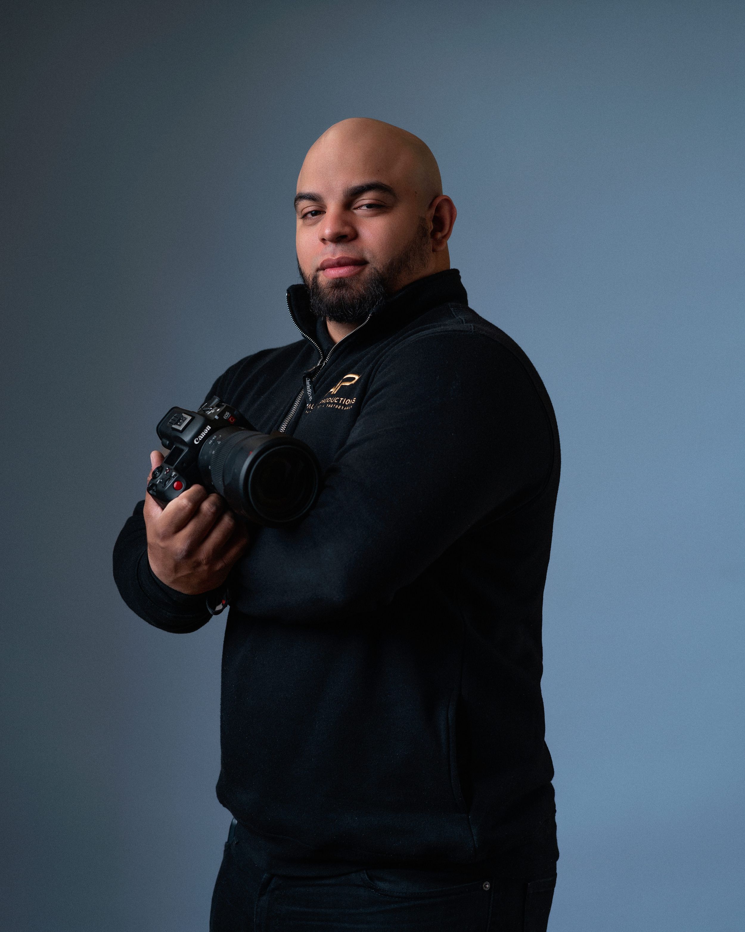 Professional photographer in black hoodie holding camera against dark background.