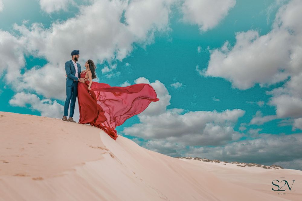 A dramatic sand dune photoshoot with flowing red dress against a vibrant turquoise sky with puffy white clouds.