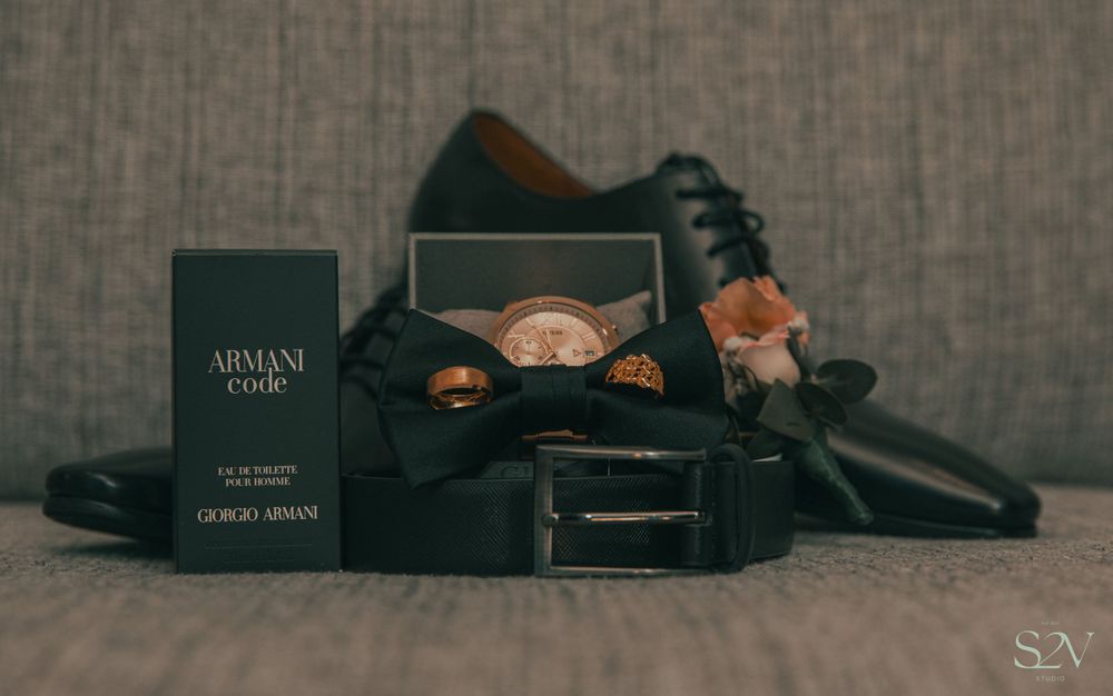 Wedding accessories including Armani cologne, dress shoes, and formal accessories arranged on grey fabric.