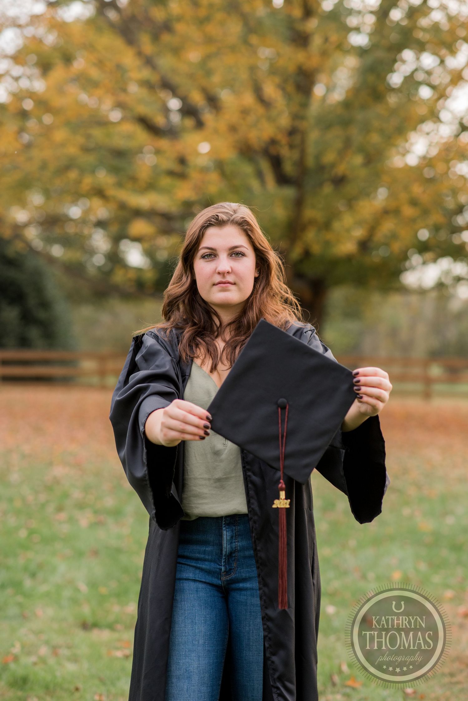 A graduate in black robes holds their cap in an outdoor fall setting with golden trees.