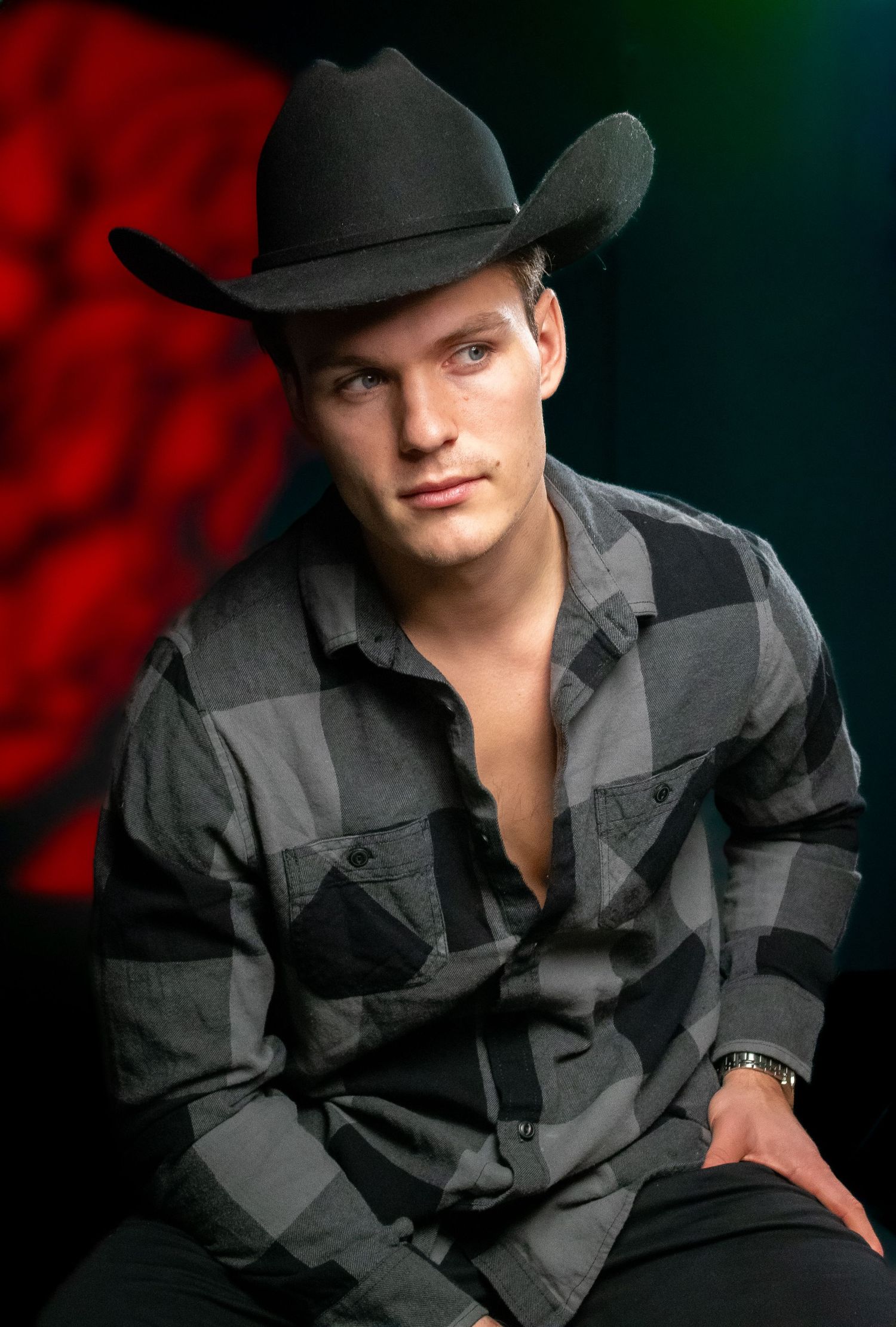 Portrait of someone wearing a black cowboy hat and plaid button-down shirt against red background.