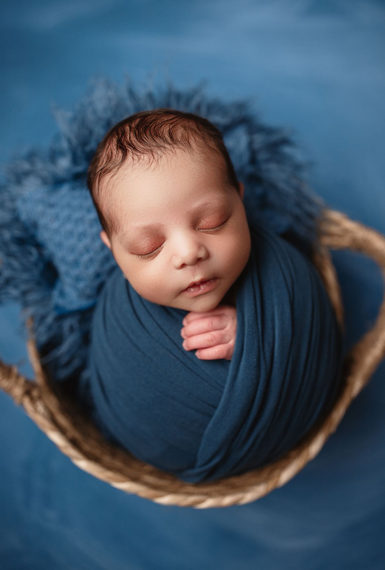 Sleeping newborn wrapped in blue fabric nestled in woven basket against blue backdrop.