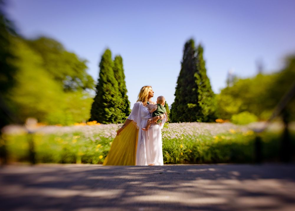 Two people in flowy dresses embrace in a sunny garden with cypress trees.