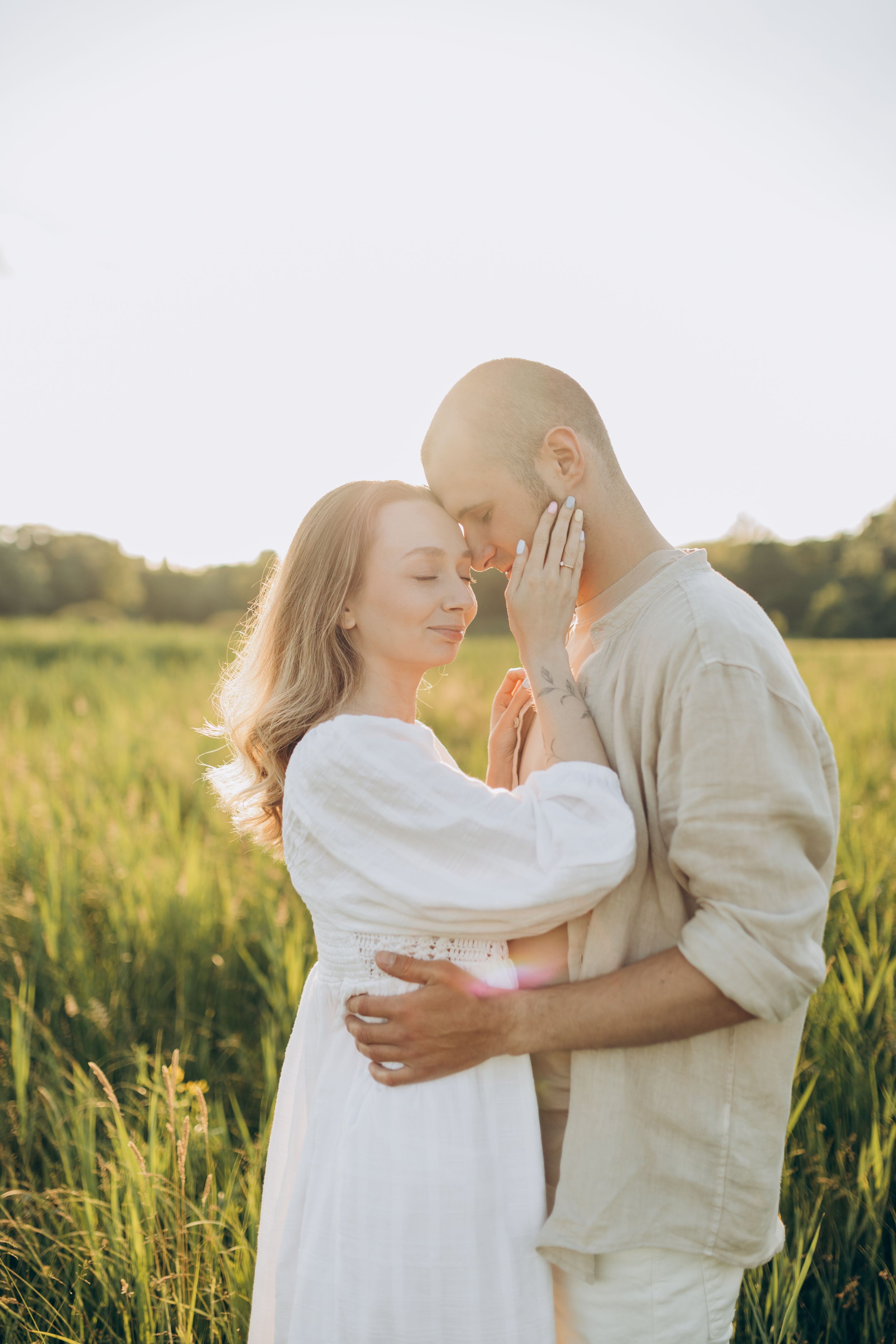 Romantic couple embraces in sunlit meadow wearing white and beige outfits.
