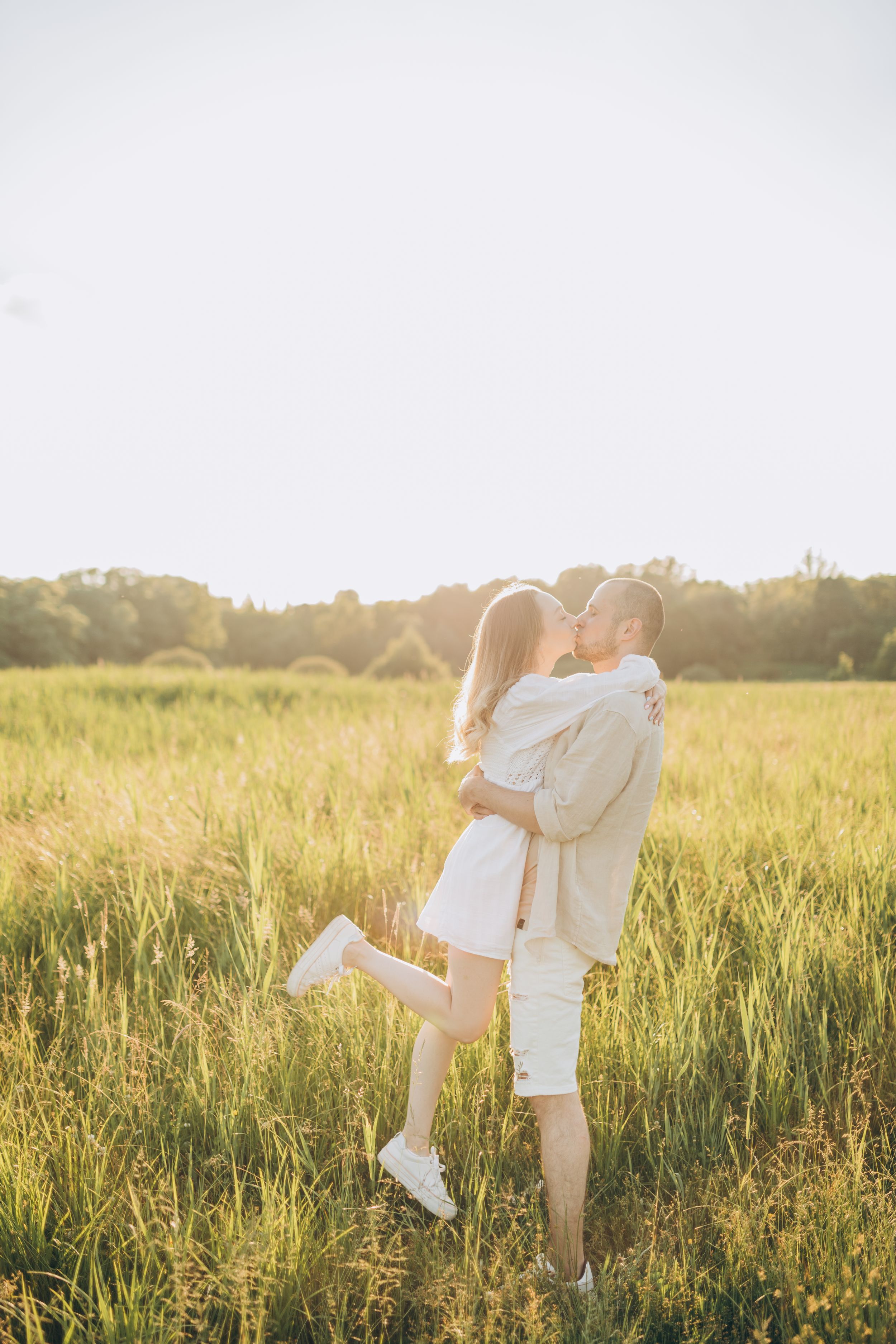 Joyful couple in white clothing embraces while standing in sunlit summer meadow.