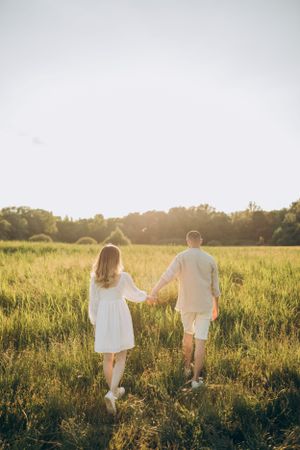 Couple walks hand in hand through golden summer meadow at sunset.