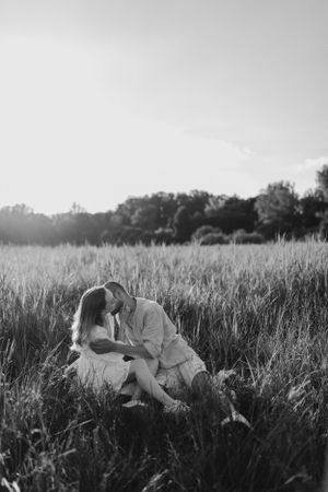 Black and white photo of couple embracing in tall grass field at sunset.