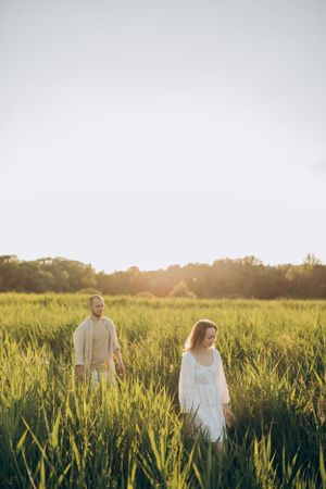 Two people in white clothing stand apart in sunlit summer meadow.