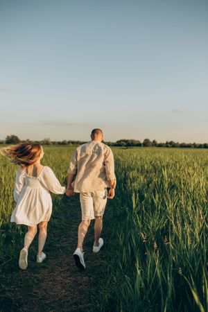 Couple in light summer clothing running along path through wheat field.