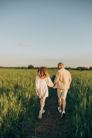 Couple holding hands while walking down path through wheat field at sunset.