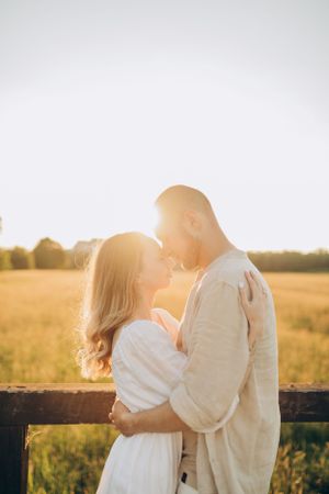 Romantic couple embraces in golden sunlit field at sunset near rustic wooden fence.